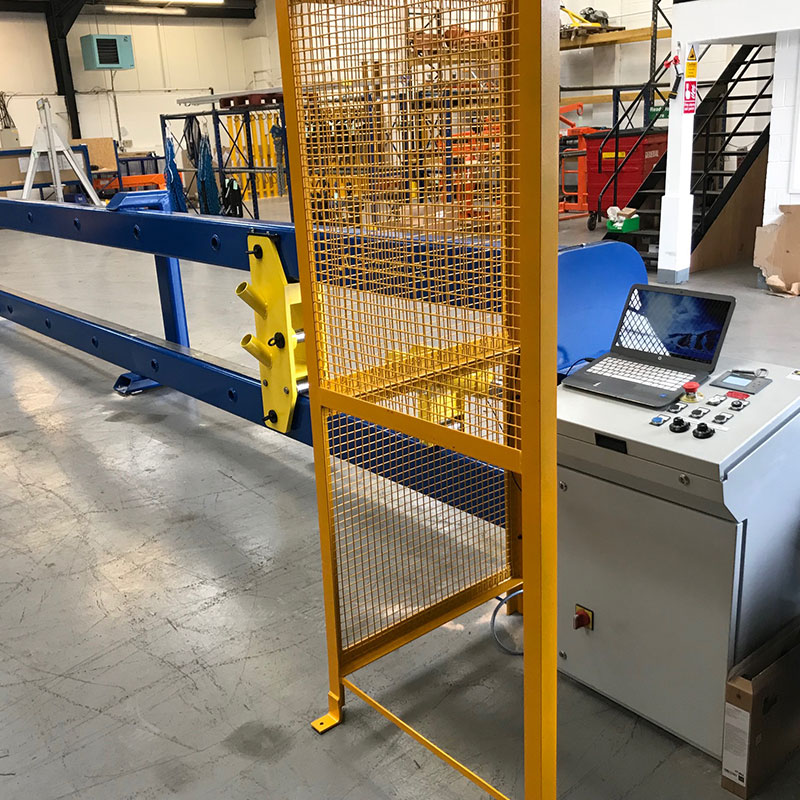 Blue and yellow tensile testing rig with control panel and mesh guard in a manufacturing facility.
